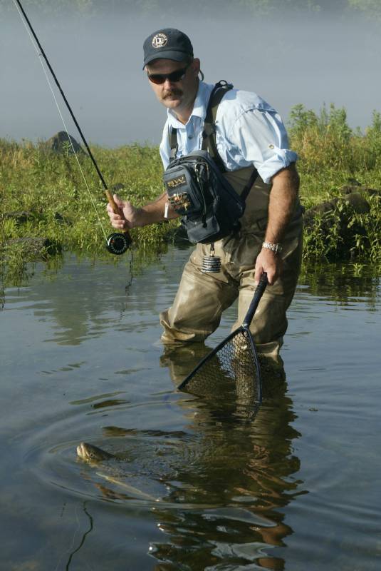 Man standing in knee-deep water, holding fly rod and netting a fish